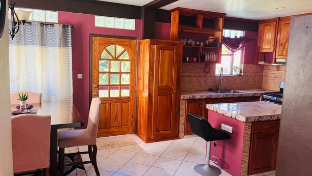 A kitchen with wooden cabinets, a dining area with a table and chairs, and a large window with curtains. - Nature's Luxurious Living (Roseau)