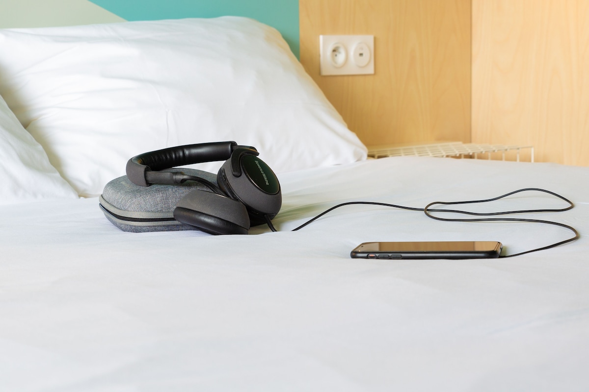 Ein Bett mit weißen Laken, einem Smartphone und einem Kopfhörerpaar darauf.