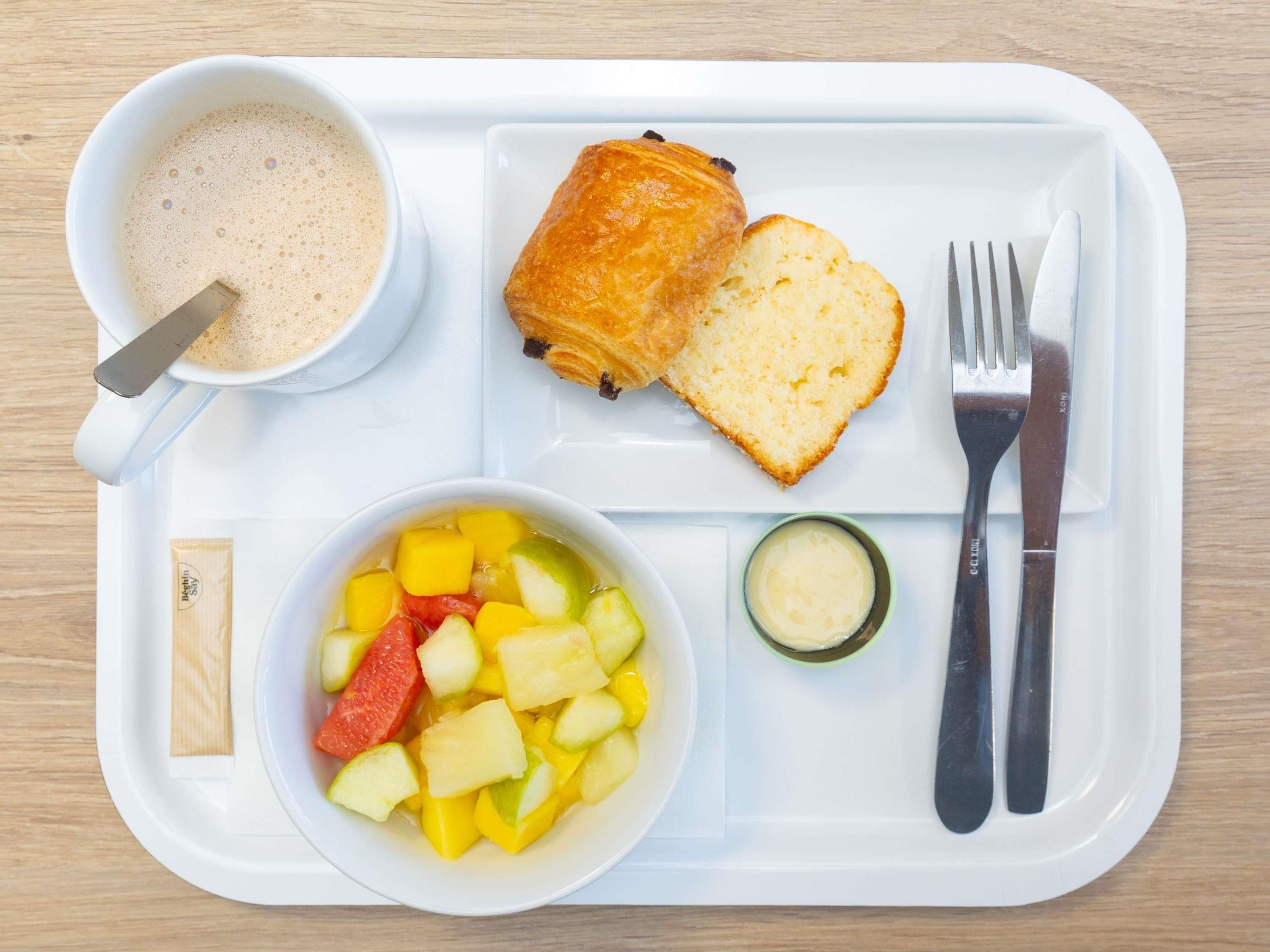 A tray with a cup of coffee, a croissant, a slice of bread, a bowl of fruit, a small container of butter, and cutlery.