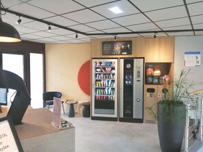 Un intérieur moderne avec des distributeurs automatiques, une grande plante en pot et un panneau indiquant une zone de service ouverte 21 heures sur 24.