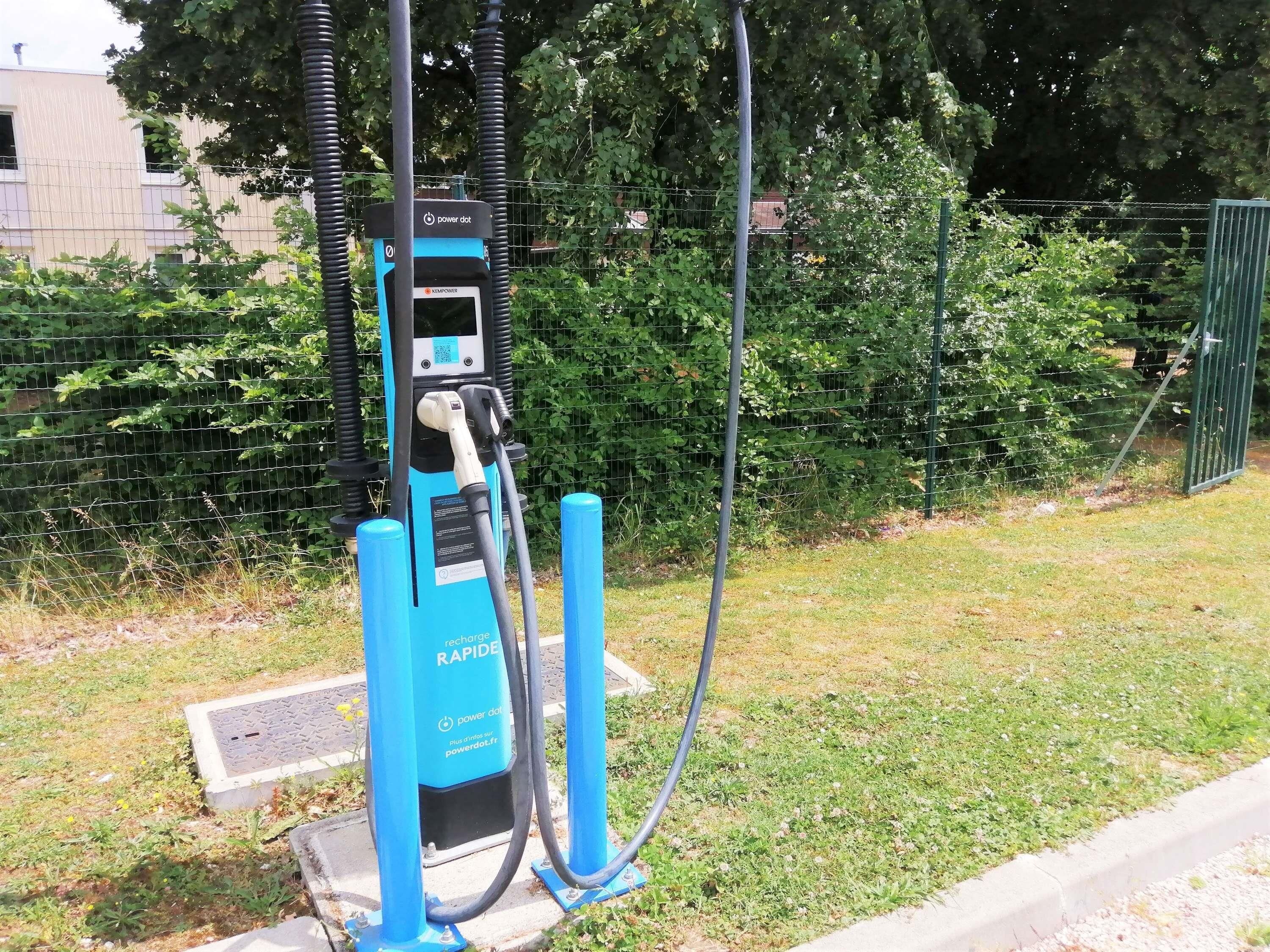 Een blauw laadstation voor elektrische voertuigen met een QR-code, buiten gelegen met een gazon en een hek op de achtergrond.