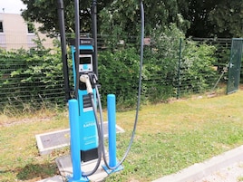 A blue electric vehicle charging station with a QR code, located outdoors with a grassy area and a fence in the background.
