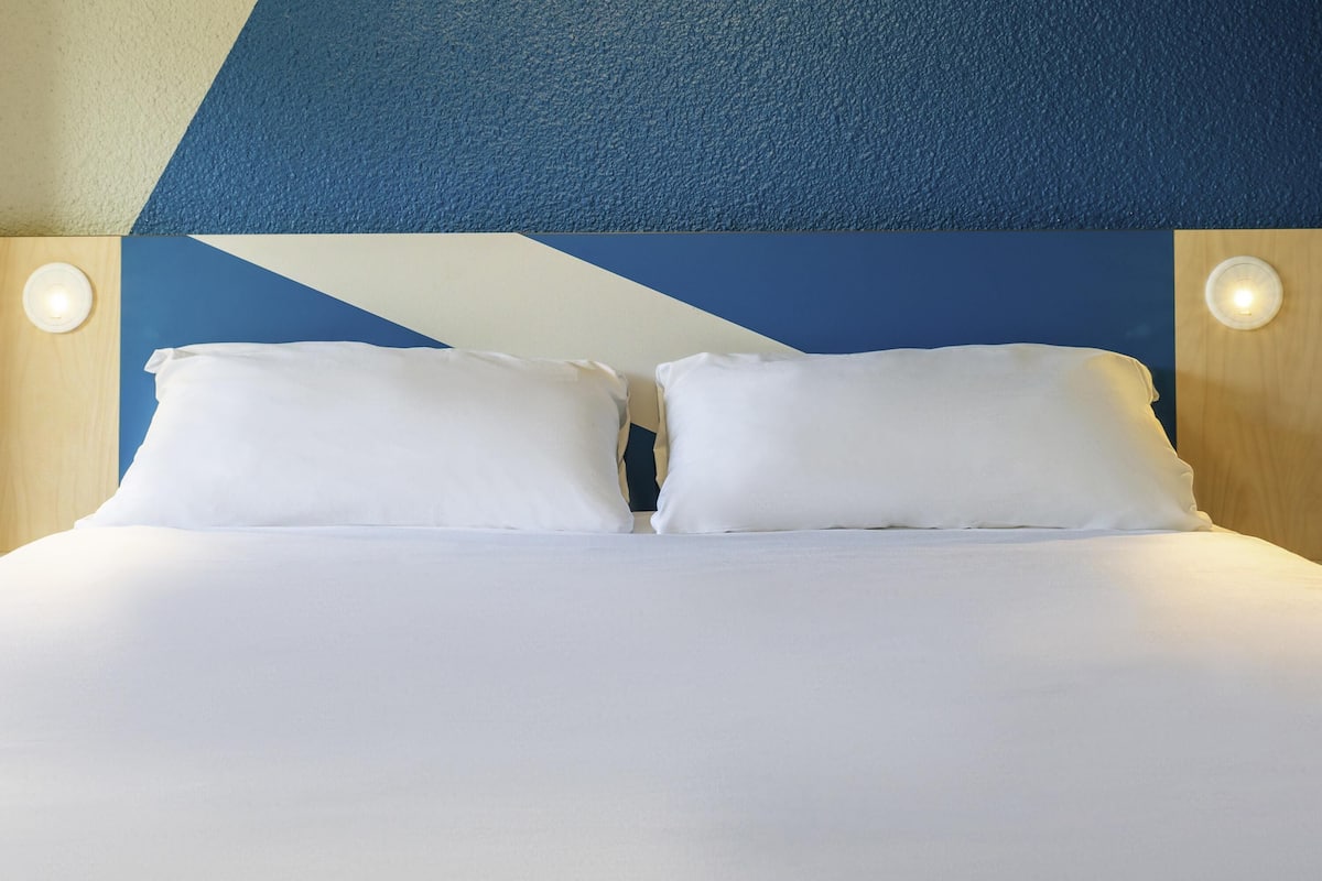 Een bed met witte lakens en twee kussens, een blauwe muur met een geometrisch patroon en twee ronde wandlampen.