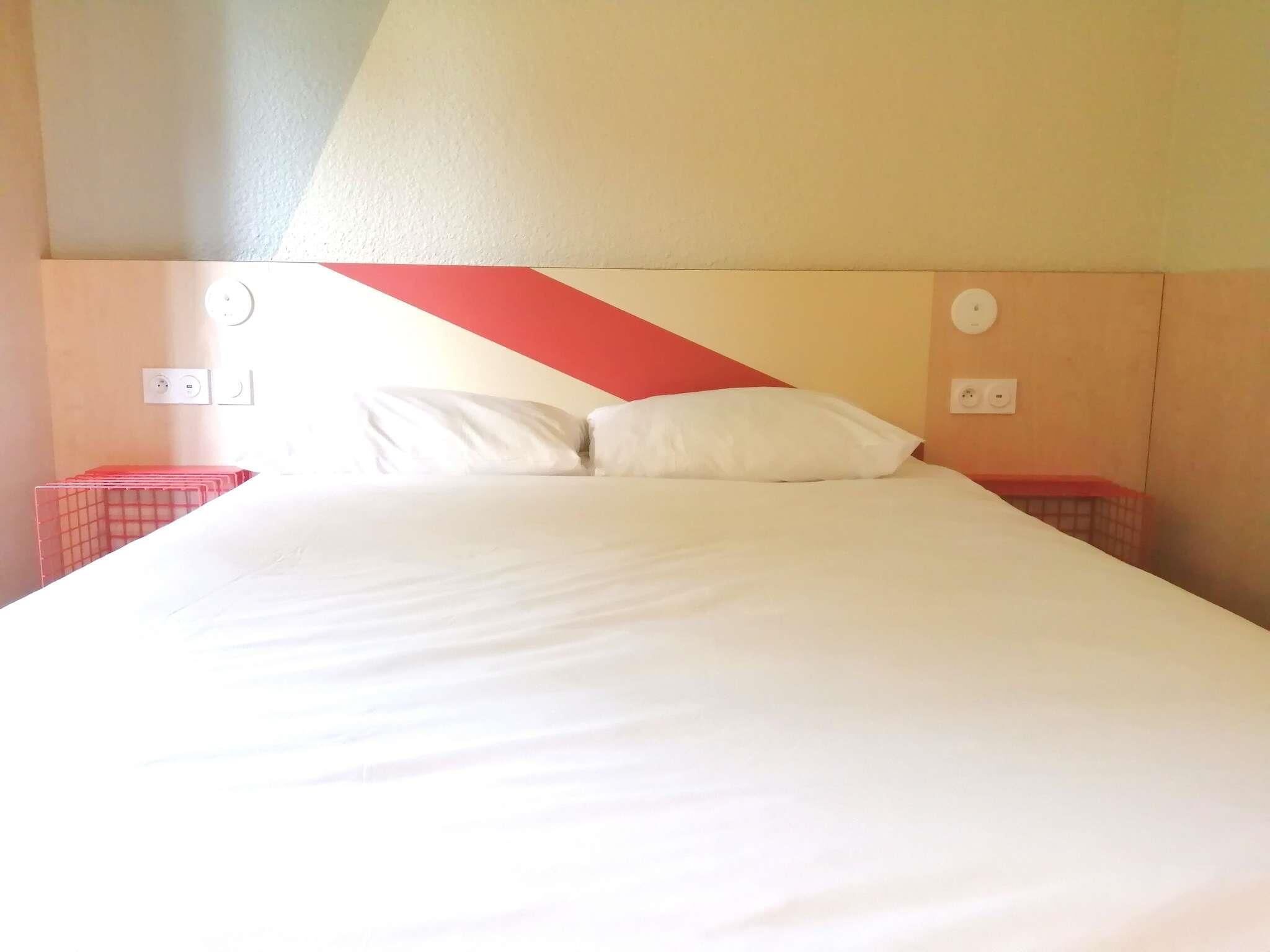Een bed met wit beddengoed, twee kussens en een hoofdbord in rood en beige. Er zijn twee nachtkastjes met rode manden van ijzerdraad en een stopcontact.