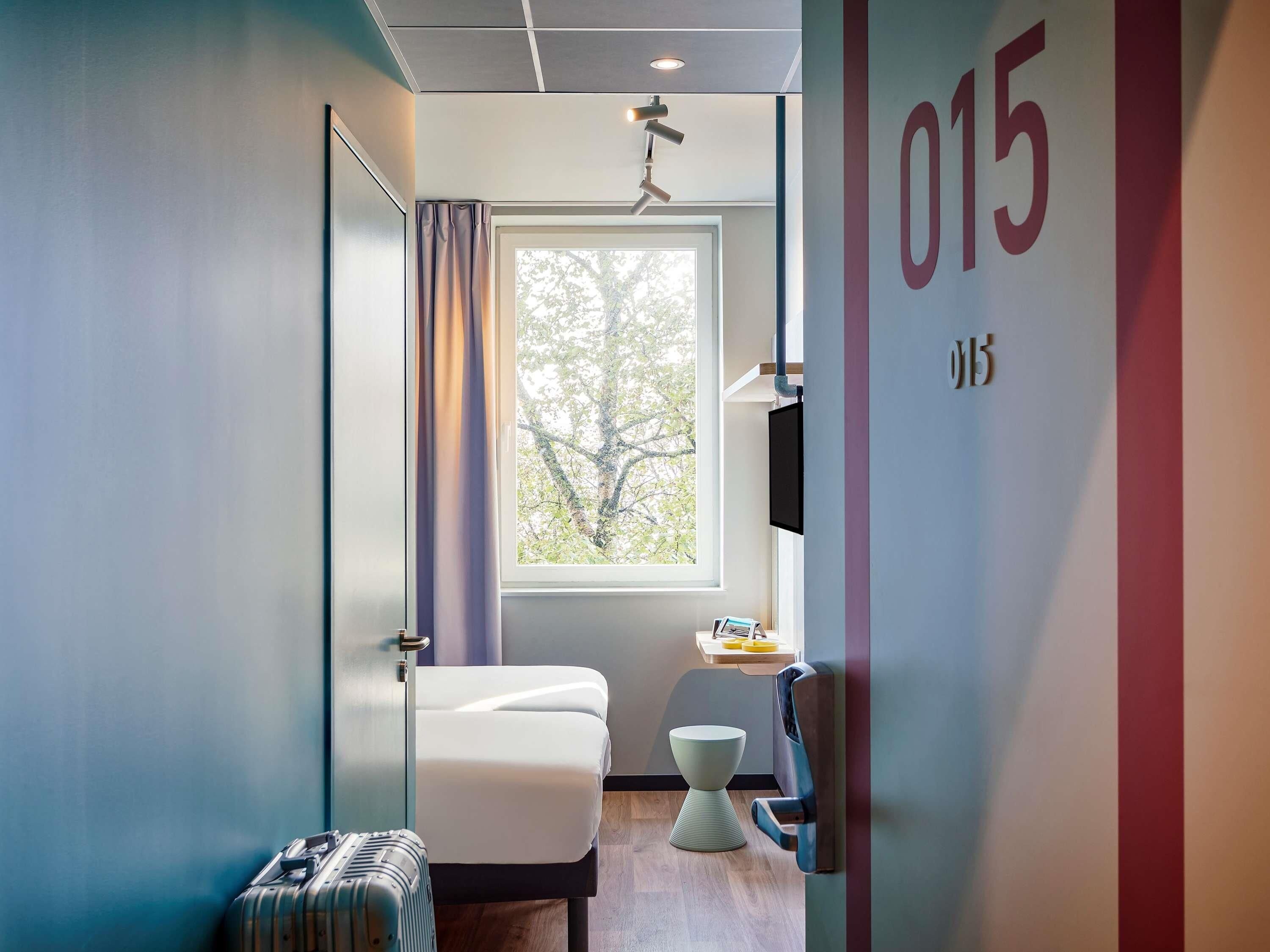 Habitación de hotel moderna con cama, inodoro, zona de ducha y ventana con vistas a árboles.