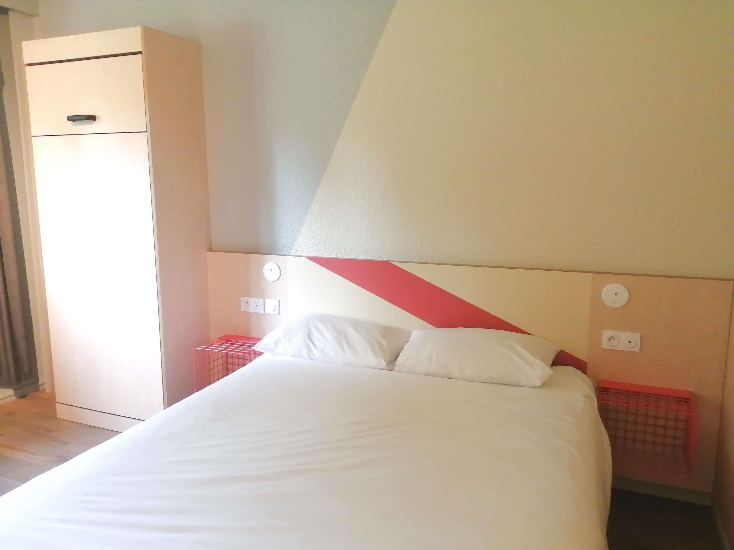 Une chambre d’hôtel avec un lit, une armoire, une tête de lit rouge et blanche et une prise de courant au mur.