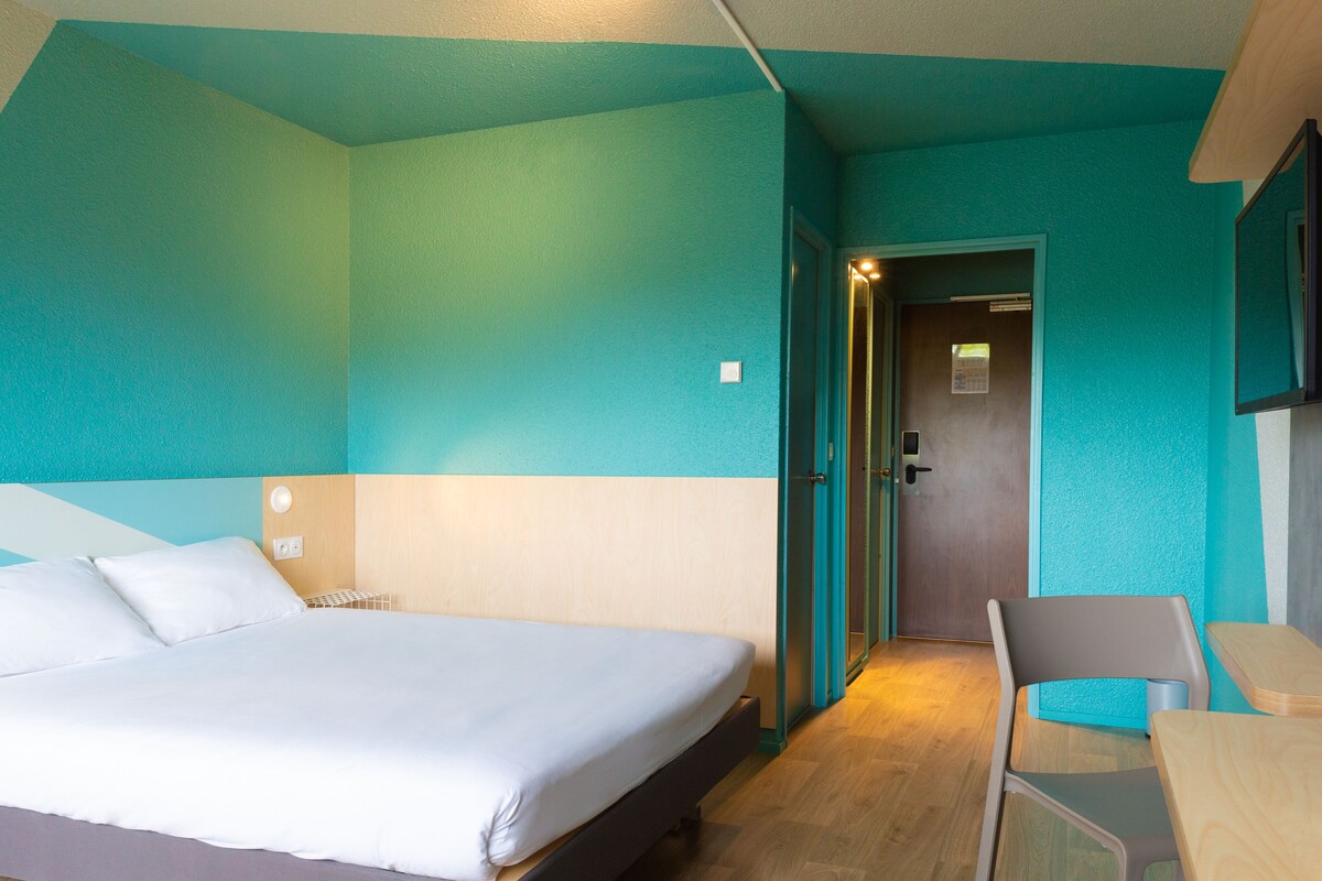 Ein Hotelzimmer mit einem Bett, einem Schreibtisch und einem Stuhl. Eine Tür führt in einen weiteren Raum.