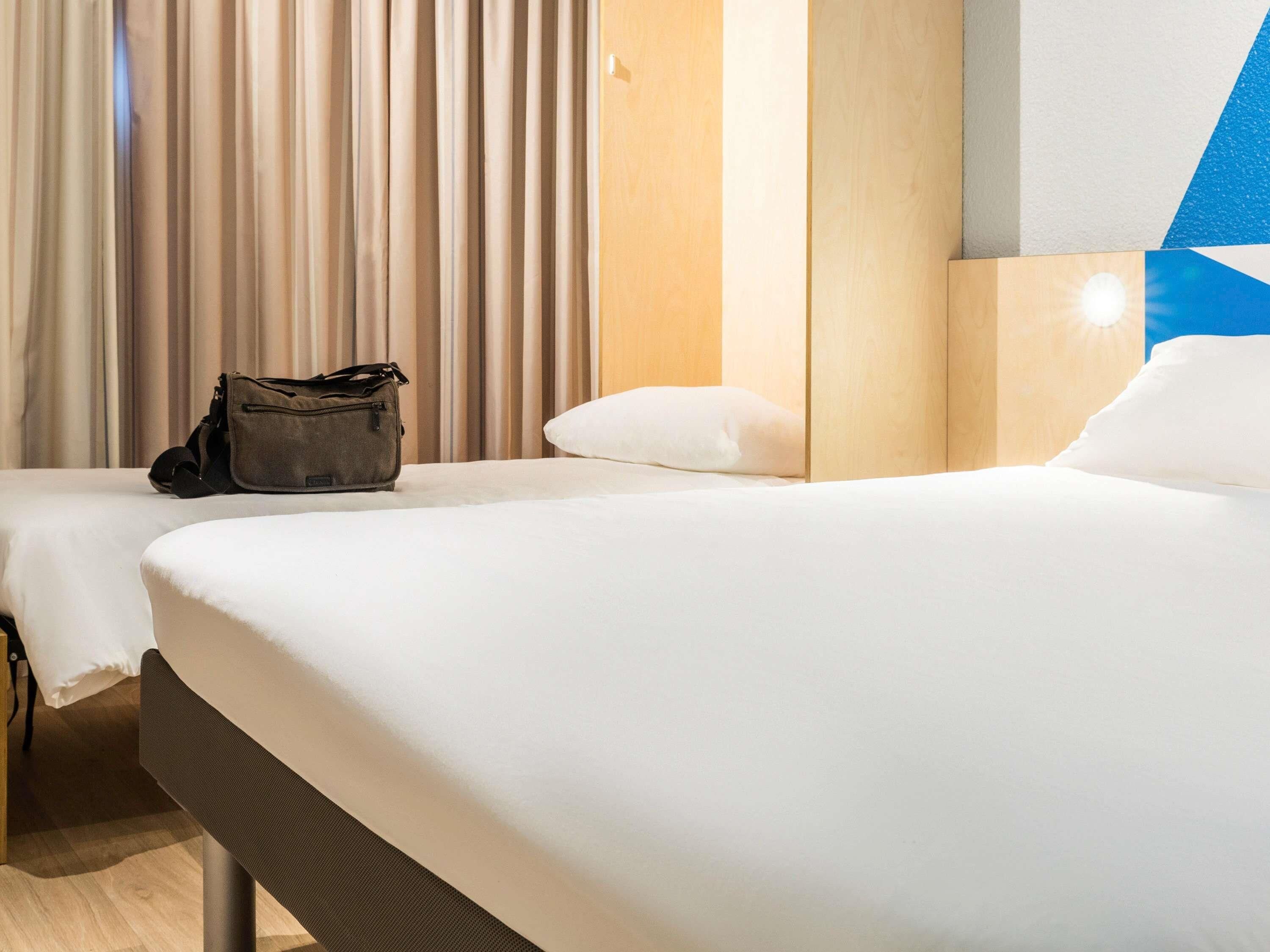 A hotel room with two beds, a wooden headboard, a beige curtain, and a small bag on one bed.