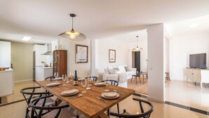 Un comedor moderno con una mesa de madera dispuesta para cuatro personas, rodeada de sillas negras. Se observa una cocina al fondo con armarios blancos y un microondas.