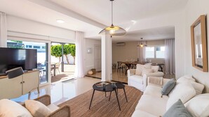 Amplia sala de estar con sofá, mesa de centro y televisor sobre un mueble. Zona de comedor con mesa y sillas, y un balcón con vistas a zonas verdes.
