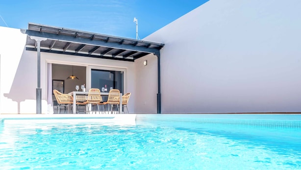 Un área de piscina moderna con una piscina azul cristalina, un espacio de descanso cubierto con sillas de mimbre y un gran ventanal corredero que da a una habitación.
