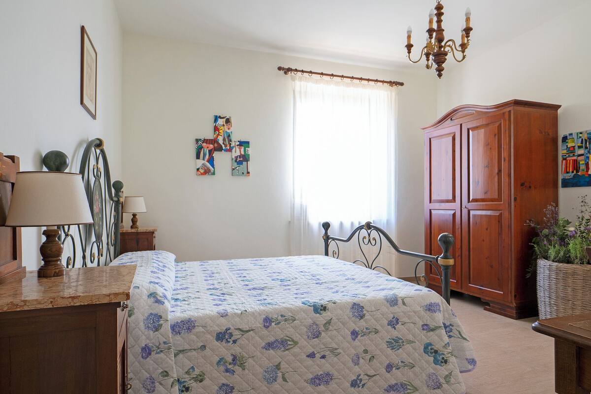 A bedroom with a bed, a wooden wardrobe, a desk with a lamp, a chandelier, and a window with curtains.
