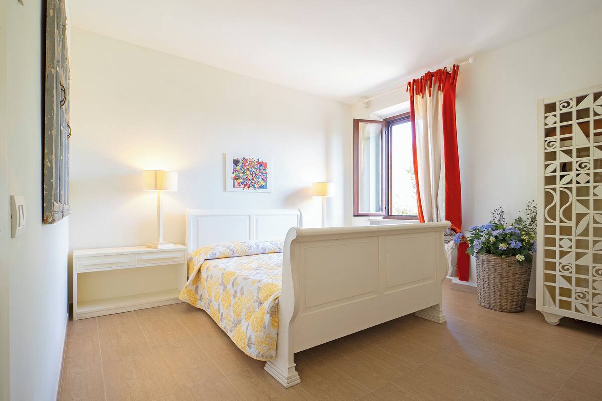 A bedroom with a bed, bedside lamps, a window with red curtains, a wall-mounted artwork, and a wicker basket with flowers.