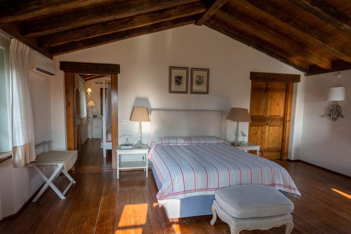 Ein Schlafzimmer mit einem großen Bett, einer Bank, zwei Nachttischen mit Lampen, Holzbalken an der Decke und einer Tür zu einem anderen Raum.