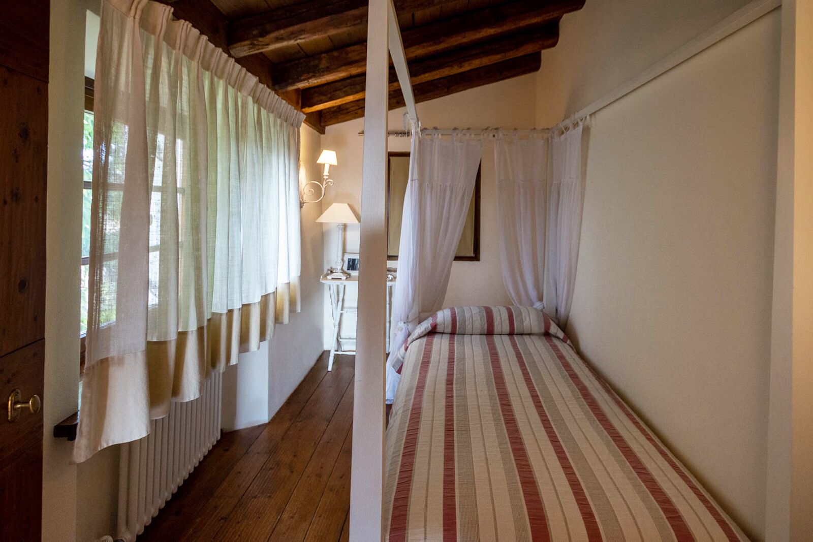 Un dormitorio con piso de madera, una cama con ropa de cama a rayas, un ventanal con cortinas translúcidas y una mesita de noche con lámpara.