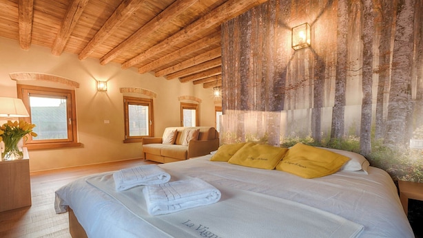 Een slaapkamer met een groot bed, houten balken in het plafond, een muurschildering met een bosmotief en een nachtkastje met een lamp.