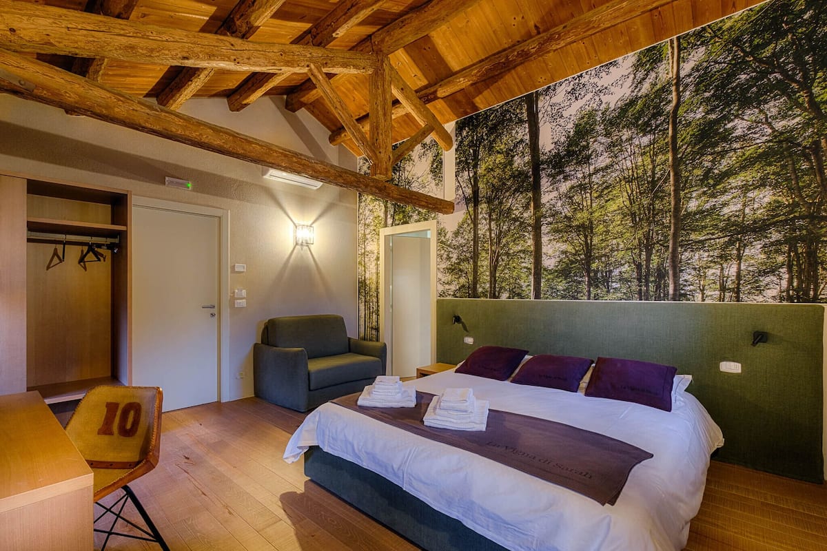 Ein Schlafzimmer mit Bett, Sessel, Tisch und Blick auf einen Wald.