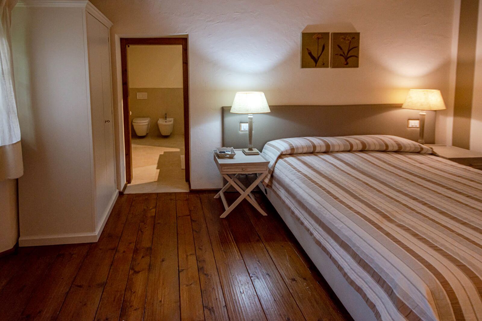 A bedroom with a wooden floor, a bed with striped bedding, a nightstand with a lamp, and a bathroom visible through an open door.
