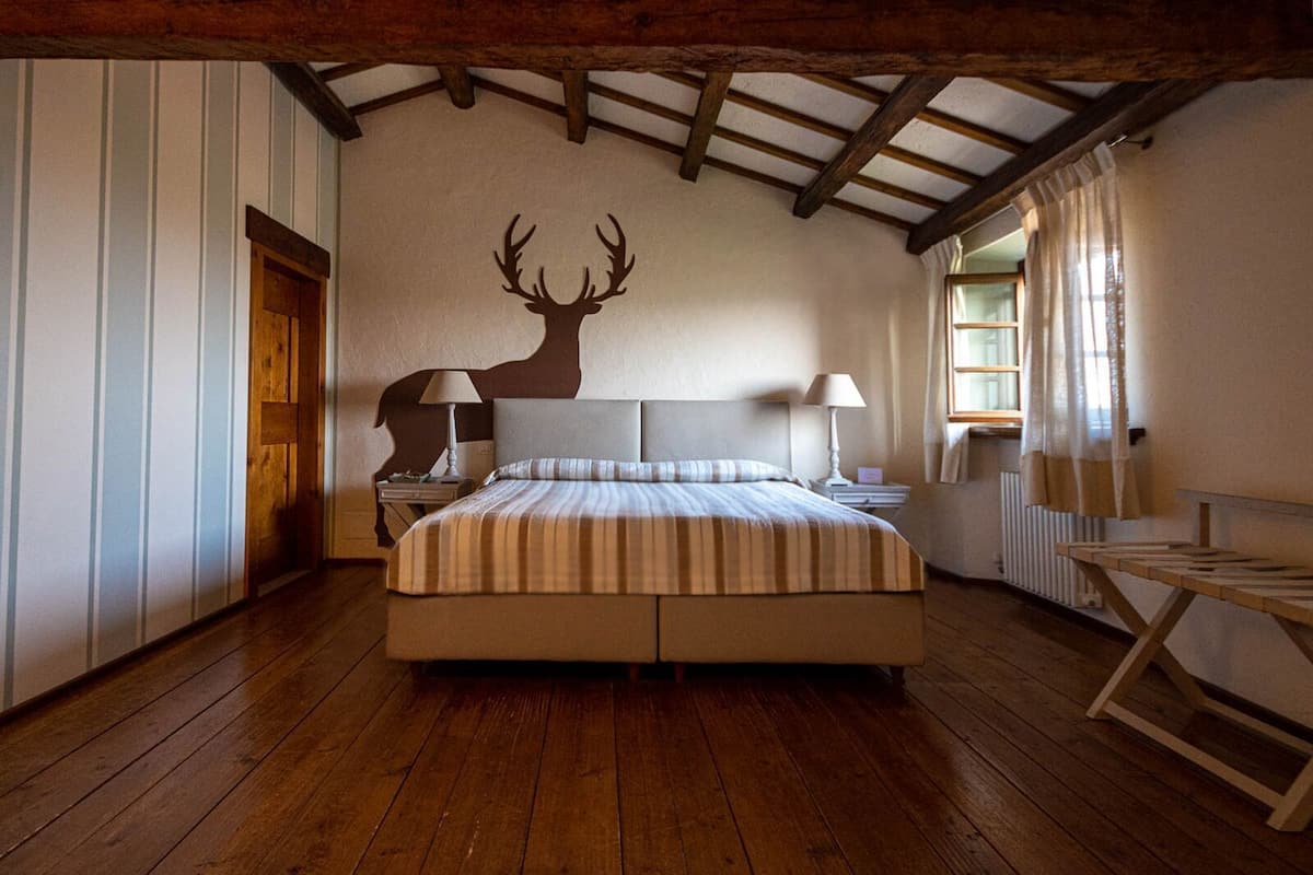 Ein Schlafzimmer mit Bett, Holzboden, einem Dekorationsobjekt in Form eines Reh-Kopfes, einem Fenster mit Vorhängen und einem Heizkörper.