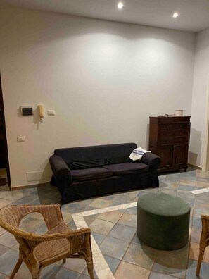 Een woonkamer met een donkergrijze bank, een rieten fauteuil, een groene poef en een houten kast.