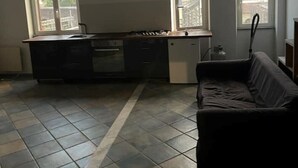 A compact kitchen with a gas stove, a built-in oven, and a small refrigerator. There are two windows providing natural light and a view of buildings outside. A dark-colored sofa is placed in the living area. - Loft SanLorenzo (Roma)