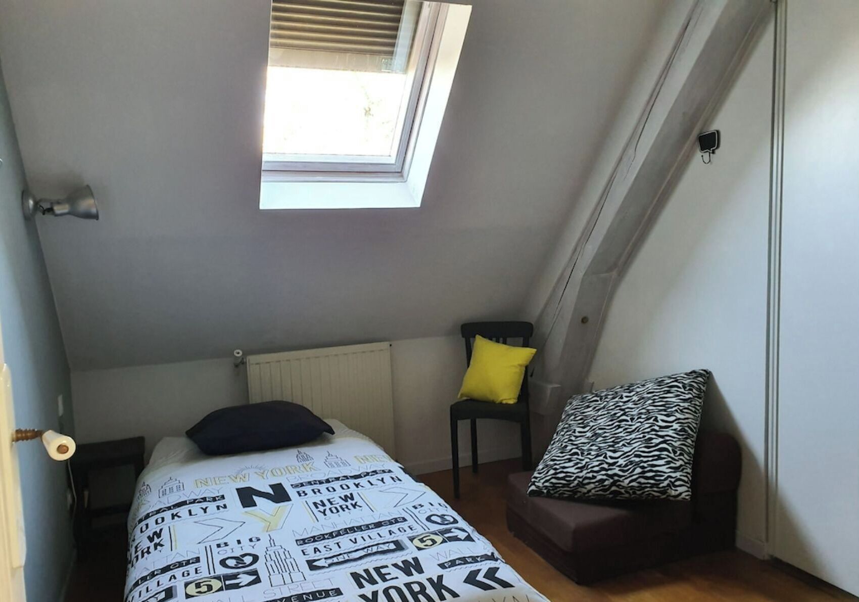 A small, narrow room with a sloped ceiling and a skylight. There is a bed with a zebra-patterned pillow and a chair with a yellow pillow.
