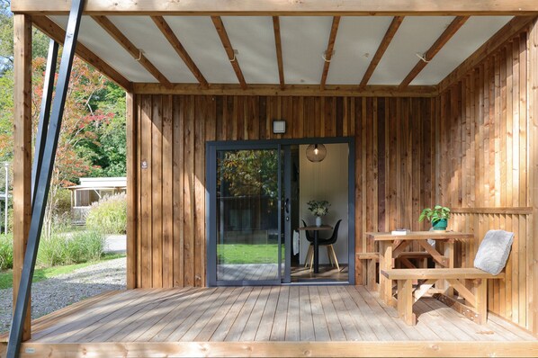 Una terraza de madera con un porche cubierto, una puerta corrediza de vidrio que da a un comedor con mesa y sillas, y una pequeña planta en maceta sobre la mesa.