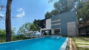 Un espace moderne aménagé au bord de la piscine, offrant une vue dégagée sur la mer, et entouré d’arbres et d’un bâtiment contemporain.