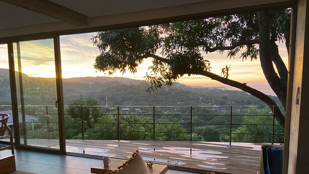Une pièce dotée d’une grande porte-fenêtre coulissante donnant sur un balcon avec vue sur un paysage vallonné au coucher du soleil.