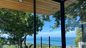 Une chambre avec un grand lit, un plafond en bois et une vue sur l’océan et la végétation.