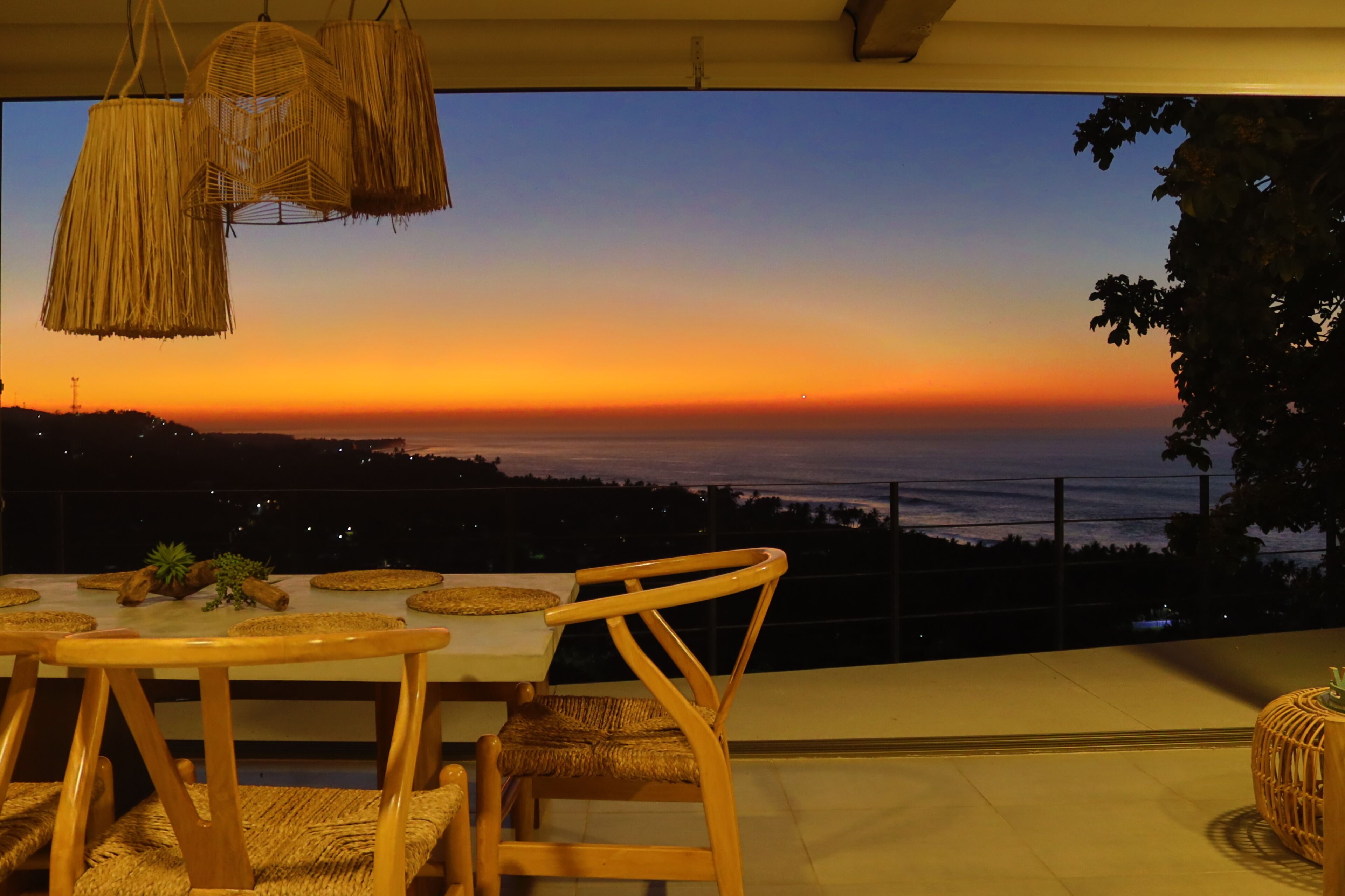 Un comedor con vista a la puesta de sol sobre el océano, con una mesa, sillas e iluminación colgante.