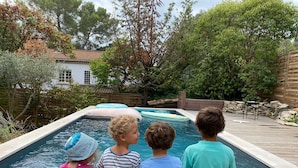 A swimming pool with four children sitting on the edge, surrounded by trees and a wooden deck.