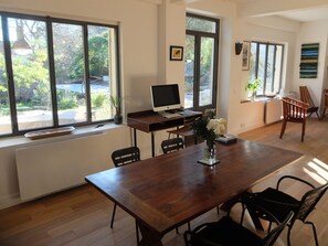Un espace repas moderne avec une table en bois, des chaises, un bureau avec un ordinateur et un écran, et de grandes fenêtres donnant sur l’extérieur.