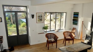 A room with a large window, wooden flooring, two wooden chairs, a bench, and a fireplace.