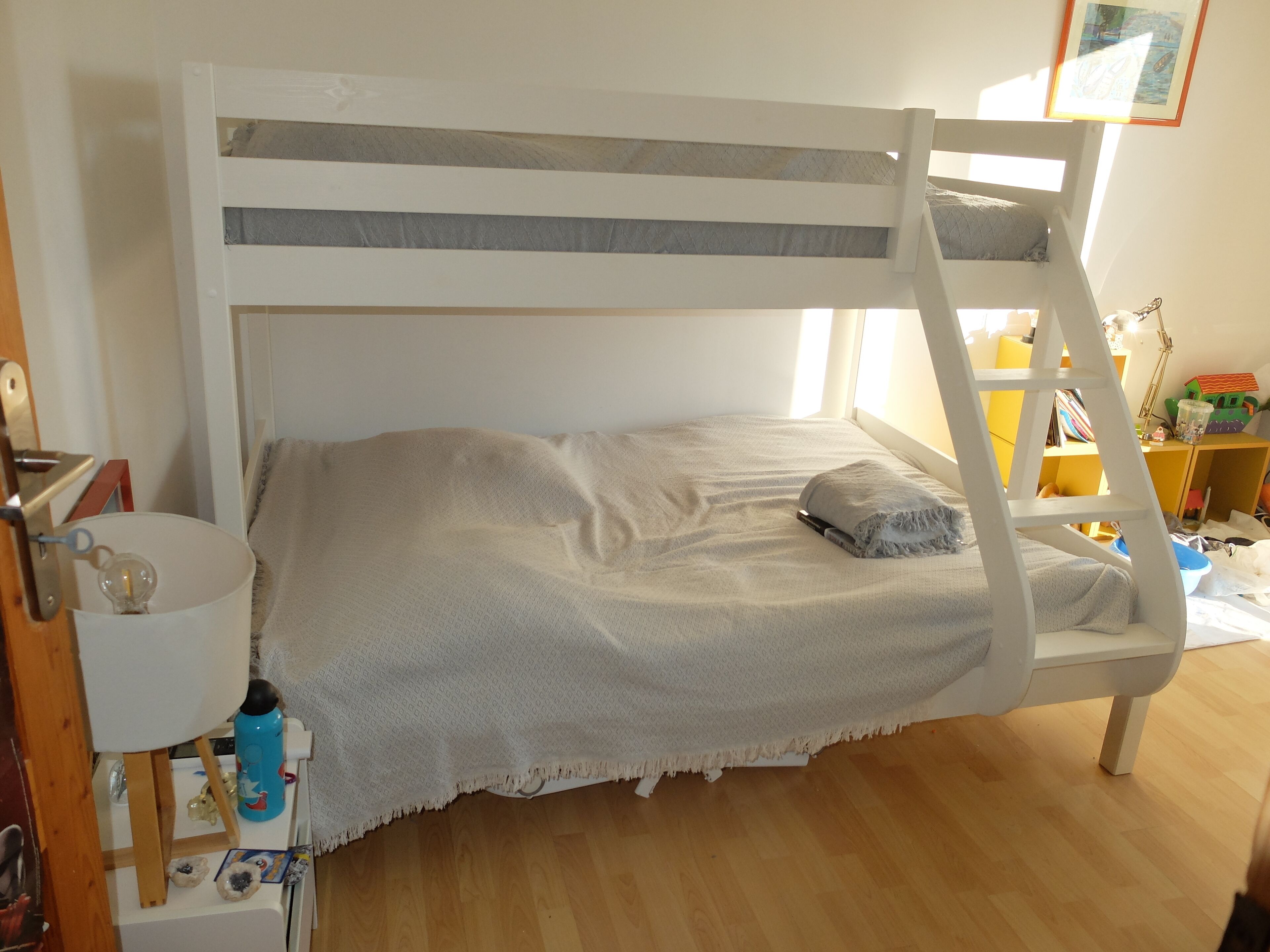 Un letto a castello con struttura bianca e testiera grigia, un comodino con una lampada e una piccola mensola con libri e giocattoli.