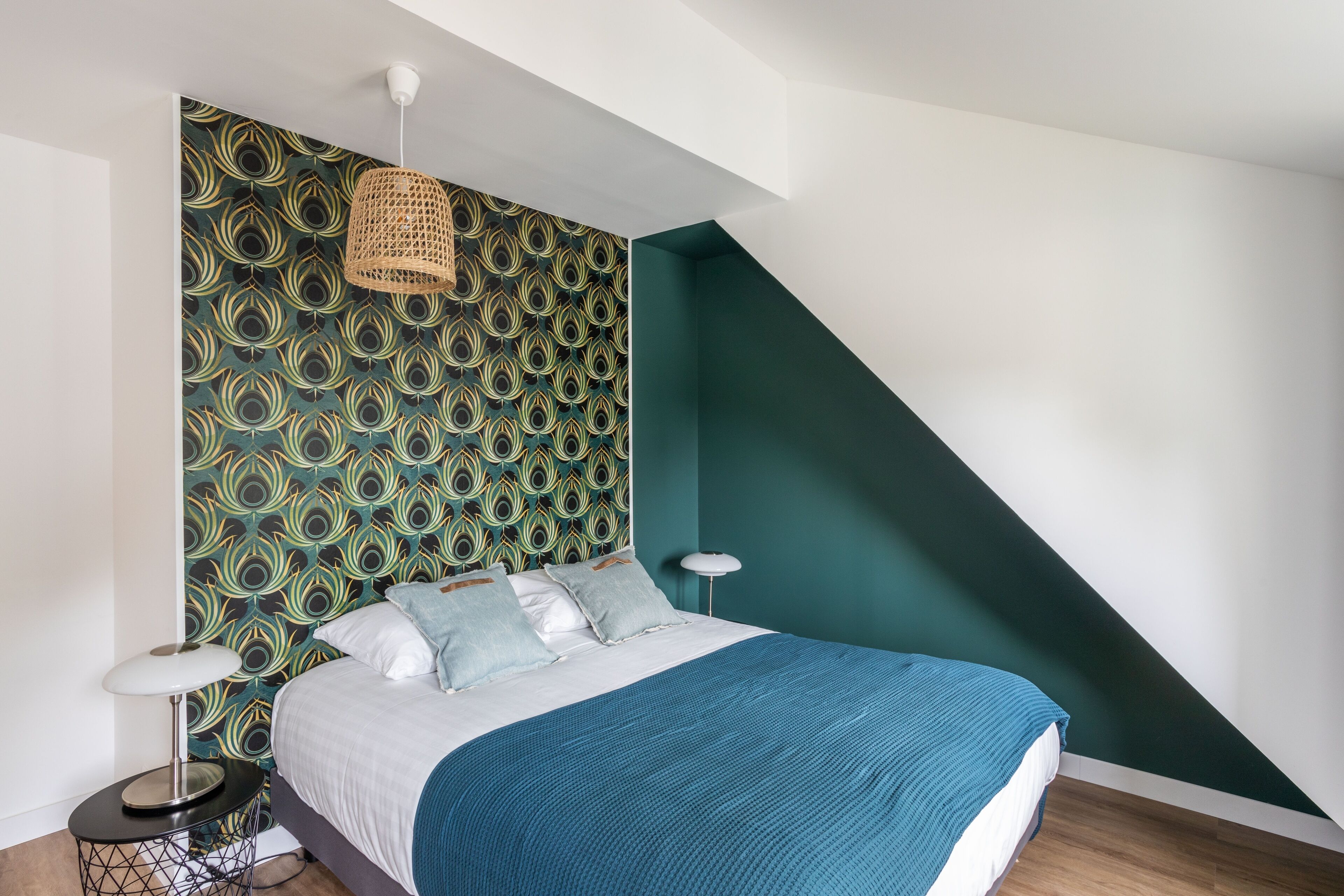 Ein Schlafzimmer mit ornamentalem Wandpapier, einem Bett mit blauer Decke und einer hängenden Leuchte.