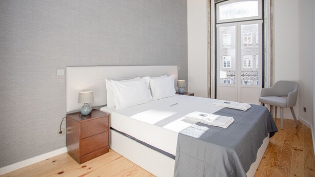 Une chambre moderne avec un grand lit, des tables de chevet, une chaise et une fenêtre donnant sur des immeubles.