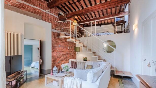 A modern living room with a white sofa, a coffee table, and a brick wall. There is a staircase with a white railing leading to an upper level. - Elegant Apartment in a Quiet Street inside the Walls Of Lucca (Lucca)