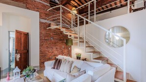A cozy living room with a white sofa, a wooden coffee table, and a brick wall. A white staircase with a round mirror is visible in the background. - Elegant Apartment in a Quiet Street inside the Walls Of Lucca (Lucca)