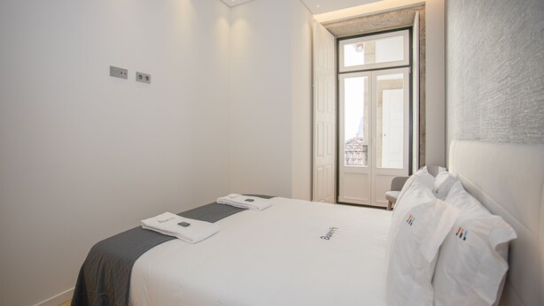 Un lit bien fait, avec une couette blanche et un plaid gris, une petite table de chevet avec une lampe, et une porte donnant sur un balcon.