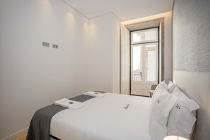Una cama bien hecha con edredón blanco y manta gris, una mesita de noche con lámpara, y una puerta que da a un balcón.