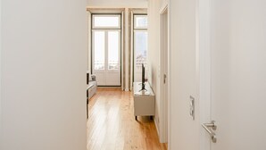 Un couloir étroit, doté d’un plancher en bois et de murs blancs, mène à une pièce avec une grande fenêtre et une porte.