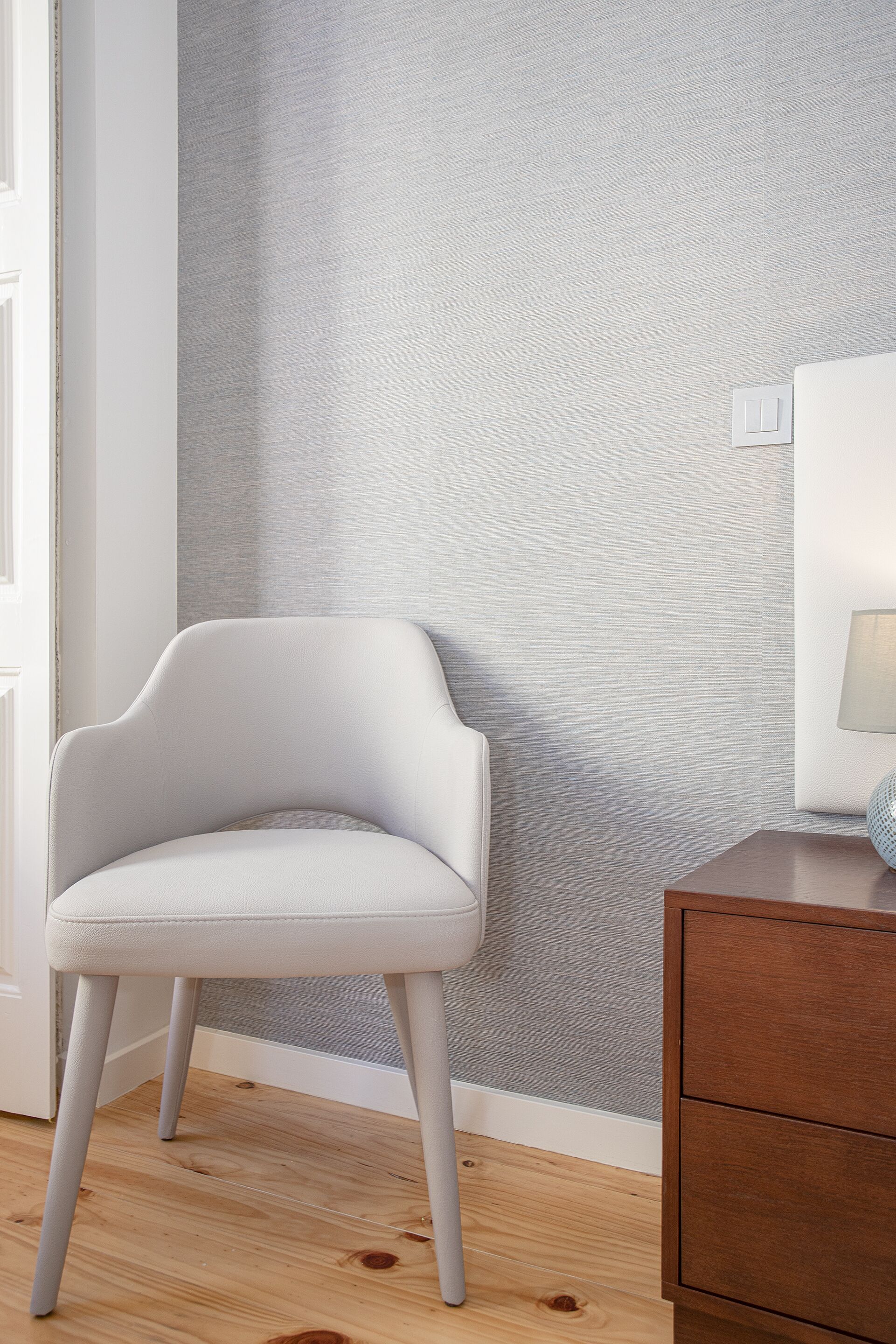 A modern room with a white upholstered chair, a wooden floor, a grey textured wall, and a wooden nightstand with a lamp.