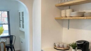 Une cuisine moderne avec des meubles de couleur sombre, un plan de travail et une machine à café. On y trouve un coin repas avec une table et des chaises, ainsi qu’une fenêtre équipée de stores.