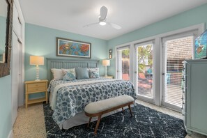 Une chambre à coucher comprenant un lit, deux tables de chevet, un banc, un ventilateur de plafond et une grande fenêtre avec des stores.