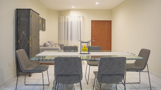 A dining area with a glass-top table, four grey upholstered chairs, a cabinet with a patterned design, a sofa, and a door. - Gaviotas Apartments (Mazatlán)