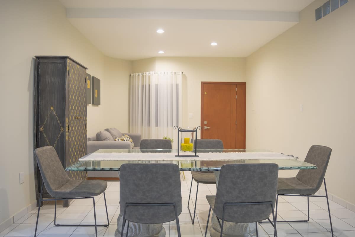 Un comedor con una mesa de vidrio, cuatro sillas grises tapizadas, un armario con diseño decorativo, un sofá y una puerta.