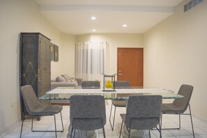 A dining area with a glass-top table, four grey upholstered chairs, a cabinet with a patterned design, a sofa, and a door.