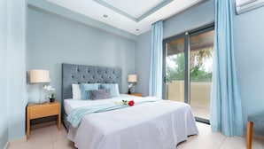 A bedroom with a large bed, two bedside tables, a nightstand, and a view of the outdoors through a sliding glass door. - Astarte Villas - Coco Palm Deluxe 3 Bedroom Suite (Ζακυνθος)