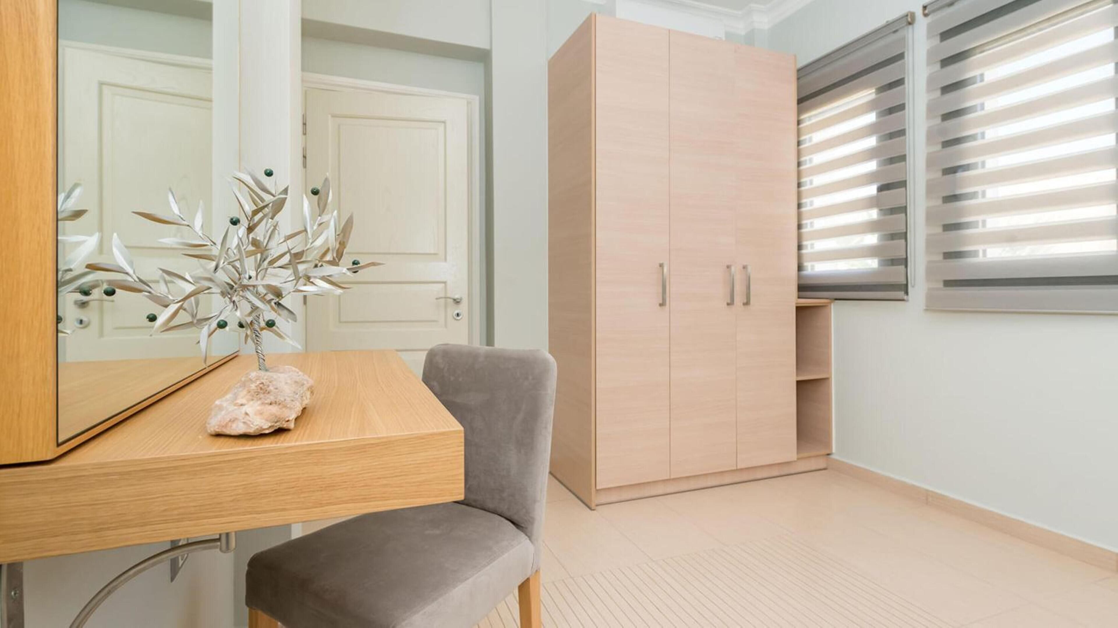 A room with a wooden desk, a grey chair, a large wardrobe, and a window with blinds.